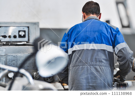 Turner in overalls works at lathe in turning shop at factory. Elderly worker turns metal part on 111138968
