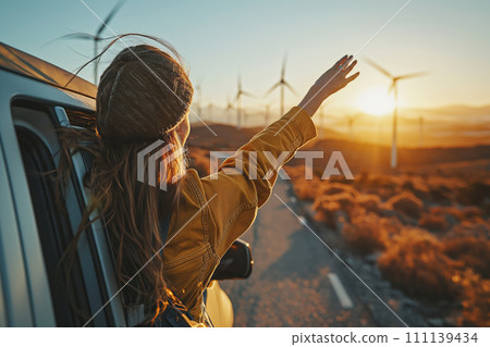 A joyful woman looks at wind generators and the sunset from a car window. 111139434