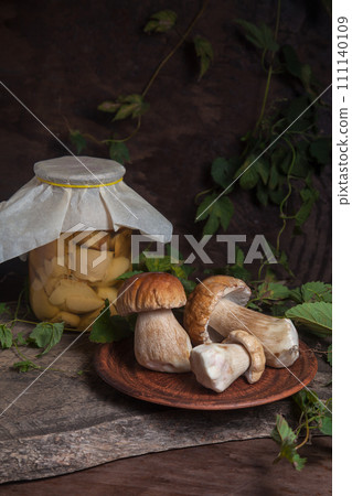 Clay plate with porcini mushroom commonly known as Boletus Edulis and glass jar with canned mushrooms on vintage wooden background.. Clay plate with porcini mushroom commonly known as Boletus Edulis and glass jar with canned mushrooms on vintage wooden background.. 111140109