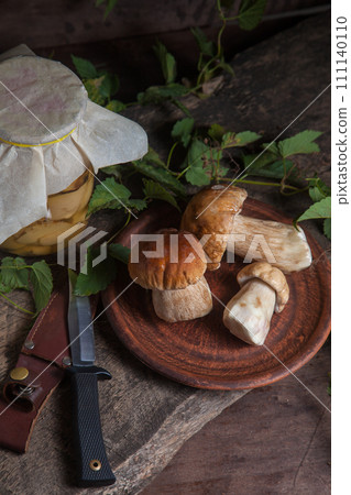 Clay plate with porcini mushroom commonly known as Boletus Edulis, glass jar with canned mushrooms and knife on vintage wooden background.. Clay plate with porcini mushroom commonly known as Boletus Edulis, glass jar with canned mushrooms and knife on vintage wooden background.. 111140110