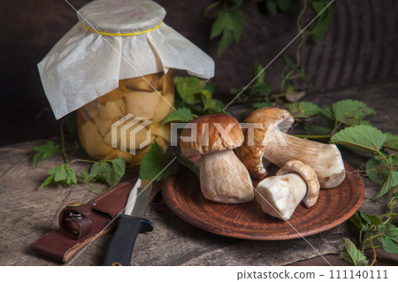 Clay plate with porcini mushroom commonly known as Boletus Edulis, glass jar with canned mushrooms and knife on vintage wooden background.. Clay plate with porcini mushroom commonly known as Boletus Edulis, glass jar with canned mushrooms and knife on vintage wooden background.. 111140111