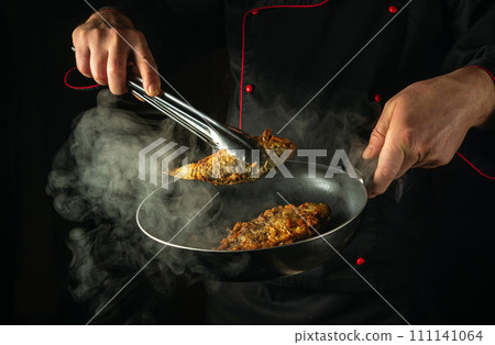 The cook is preparing fish in the kitchen. The concept of preparing a fish dish for breakfast. The chef hand uses tongs to remove hot fried carp from the frying pan 111141064