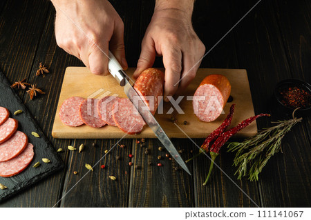 Cooking a hearty breakfast with smoked sausage by the hands of a cook on the kitchen table. Cutting sausage with a knife for a serving board 111141067