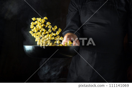 The chef is preparing a pasta dinner in a frying pan. Healthy vegetarian food and dishes recipe. Free advertising space on a dark background The chef is preparing a pasta dinner in a frying pan. Healthy vegetarian food and dishes recipe. Free advertising space on a dark background 111141069