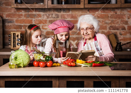 Granny with two granddaughters reading recipe from cooking book and cooking salad 111141778