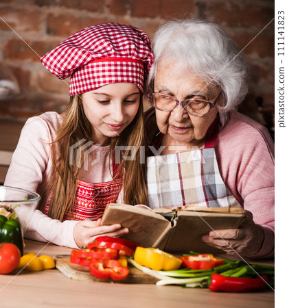 Little granddaughter reading recipe book with her granny sitting at kitchen 111141823