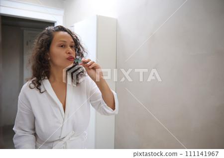 Young woman brushing teeth, looking at her mirror reflection in the home bathroom, dressed in white waffle bathrobe Young woman brushing teeth, looking at her mirror reflection in the home bathroom, dressed in white waffle bathrobe 111141967