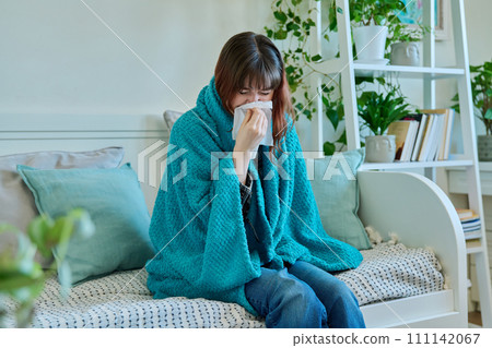 Young sick woman sitting under warm blanket sneezing coughing in handkerchief Young sick woman sitting under warm blanket sneezing coughing in handkerchief 111142067