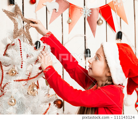 little girl in red hat putting a star on top of christmas tree in decorated room 111143124