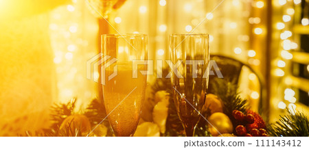 Two champagne glasses filled with champagne are placed on a table with a fruit arrangement. The glasses are surrounded by lights, creating a festive atmosphere. 111143412