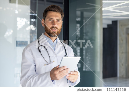 Portrait of a serious young male doctor standing in a white coat in the office of the clinic, holding a tablet in his hands and confidently looking at the camera. 111145236
