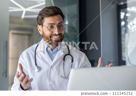 Close-up photo of a young smiling specialist doctor dressed in a robe sitting in a hospital office in front of a laptop and talking online via video call, consulting a patient remotely. 111145252