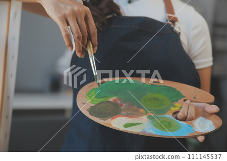 Cropped image of female artist standing in front of an easel and dipping brush into color palette 111145537