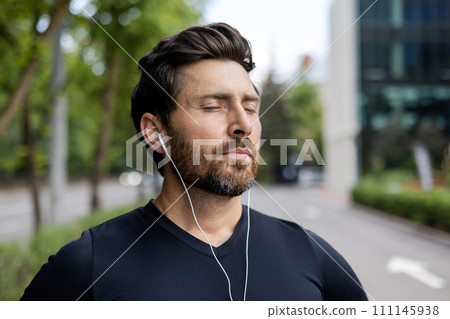 Close-up photo of a young male athlete wearing headphones and black sportswear standing outside on the street, listening to music and resting with his eyes closed. Close-up photo of a young male athlete wearing headphones and black sportswear standing outside on the street, listening to music and resting with his eyes closed. 111145938