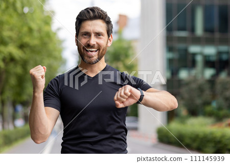 Close-up portrait of young happy male sportsman rejoicing in success showing victory gesture with hand, satisfied with result on smart watch and smiling at camera. Close-up portrait of young happy male sportsman rejoicing in success showing victory gesture with hand, satisfied with result on smart watch and smiling at camera. 111145939