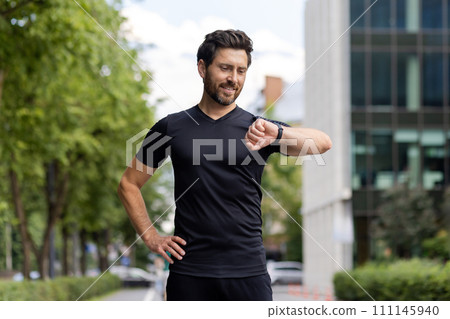 A sporty smiling young man standing on a city street checks the time after training on a smart watch on his wrist. A sporty smiling young man standing on a city street checks the time after training on a smart watch on his wrist. 111145940