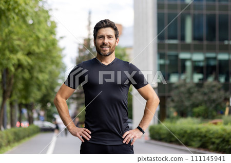 Portrait of a young man in sportswear standing outside on the street, hands on waist, confidently and smilingly looking at the camera. Portrait of a young man in sportswear standing outside on the street, hands on waist, confidently and smilingly looking at the camera. 111145941