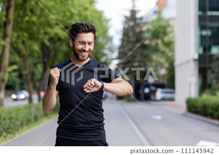 Happy young man doing sports and running, standing on city street, looking at wrist tracker and showing success gesture with hand. Happy young man doing sports and running, standing on city street, looking at wrist tracker and showing success gesture with hand. 111145942