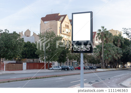 Urban digital clock post with a blank advertising panel against a cityscape, ideal for promotions. Vertical billboard mockup, empty display. Copy space for text, picture. 111146363