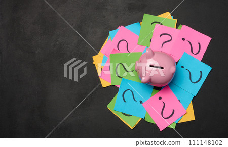 Ceramic piggy bank and stickers with drawn question marks on a black background, top view Ceramic piggy bank and stickers with drawn question marks on a black background, top view 111148102