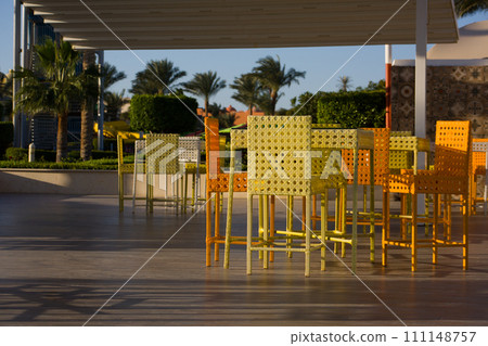 Cute summer cafe with colorful wicker chairs and a table against the backdrop of an exotic park with palm trees on a sunny day Cute summer cafe with colorful wicker chairs and a table against the backdrop of an exotic park with palm trees on a sunny day 111148757