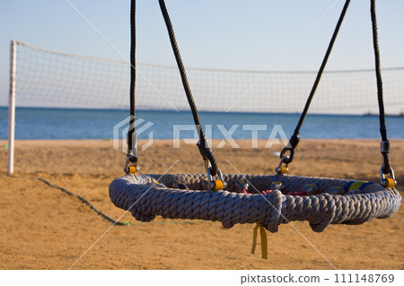Swing nest on a sandy beach against the background of a volleyball court with a net, an empty swing without people on the seashore Swing nest on a sandy beach against the background of a volleyball court with a net, an empty swing without people on the seashore 111148769
