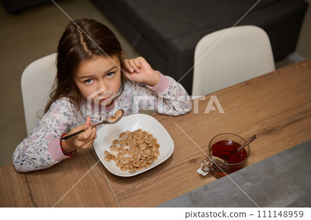 A little child girl in stylish pajamas, sitting at kitchen table and eating her healthy breakfast in the morning A little child girl in stylish pajamas, sitting at kitchen table and eating her healthy breakfast in the morning 111148959