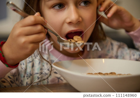 Close-up face portrait of hungry child girl holding full table spoon of sweet delicious corn oat flakes with milk honey Close-up face portrait of hungry child girl holding full table spoon of sweet delicious corn oat flakes with milk honey 111148960