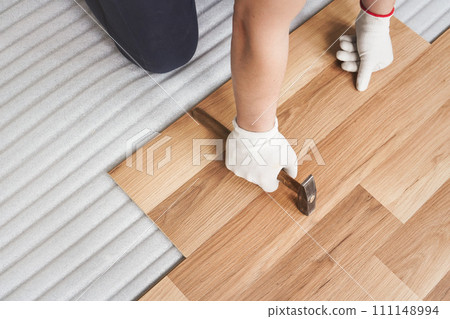 Installing laminated floor, detail on man hands holding in white gloves, hammer in one hand, leaning over wooden tiles, white foam base layer under Installing laminated floor, detail on man hands holding in white gloves, hammer in one hand, leaning over wooden tiles, white foam base layer under 111148994
