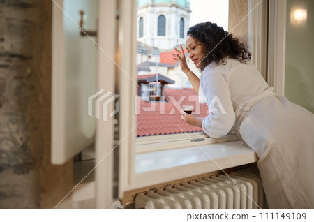 Smiling young woman greeting someone with her hand while looking out the window and enjoying her morning coffee 111149109