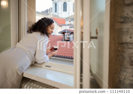 Happy female tourist looking out the window and enjoying her morning coffee, admiring a beautiful city during her travel Happy female tourist looking out the window and enjoying her morning coffee, admiring a beautiful city during her travel 111149110