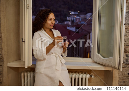 Smiling young woman in white robe, enjoying her morning espresso coffee, standing by opened window at home 111149114