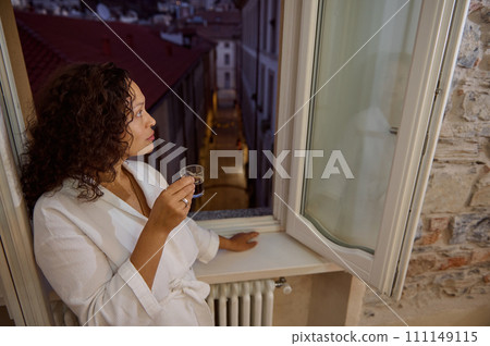 Young woman in white robe, standing at opened window, drinking her morning coffee and looking outside, enjoys of rest Young woman in white robe, standing at opened window, drinking her morning coffee and looking outside, enjoys of rest 111149115