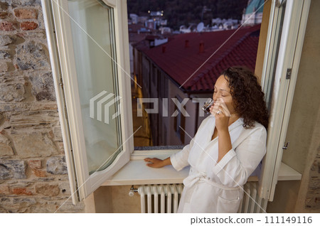 Overhead view of a sleepy woman drinking coffee, standing by window at dawn in the cozy home interior 111149116