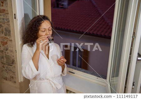Delightful young sleepy woman enjoying her morning coffee, standing by open window at dawn, overlooking the city of Como 111149117