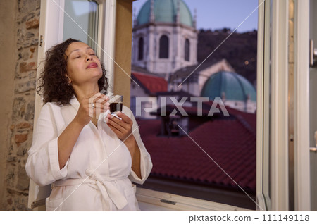 Delightful young adult woman enjoying the aroma of freshly brewed espresso coffee, standing by open window at dawn 111149118