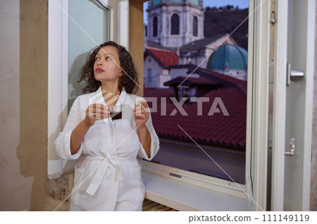 Attractive young woman holding cup of aroma espresso coffee, standing by open window overlooking the medieval cathedral 111149119