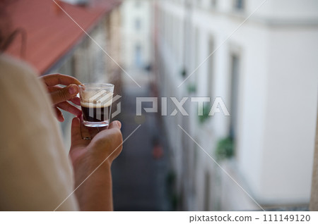 Closeup of woman's hands hold cup of freshly brewed espresso, standing by windows and admiring the beautiful city view 111149120
