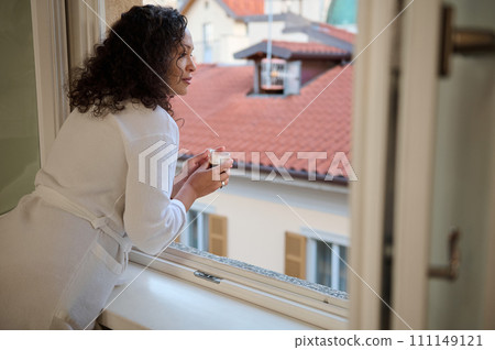 Delightful young woman in white bathrobe, looks out the window, holds a cup of coffee and enjoys the beautiful city view 111149121