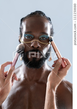 Bearded gay man applying makeup with brushes held 111150080