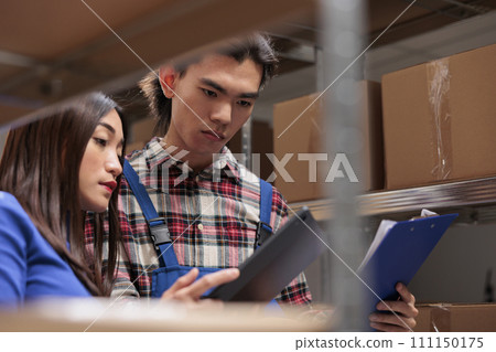 Storehouse asian workers comparing inventory data reports in storage room. Postal warehouse employees doing logistics and distribution management together near cardboard boxes shelf 111150175