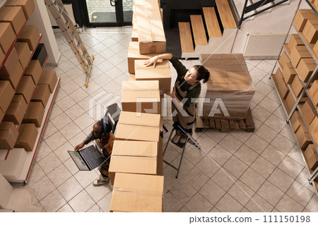 Top view of multi ethnic colleagues working in storehouse, preparing customers orders before start delivery merchandise. Storage room team in protective overall working with cardboard boxes 111150198