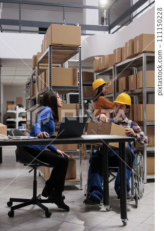 Delivery manager and coworker in wheelchair preparing parcels for shipping while managing orders from laptop at a warehouse desk. Storehouse worker with disability giving packed box to colleague 111150228