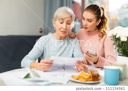 Aged woman reading documents while adult daughter making copy with phone 111154561