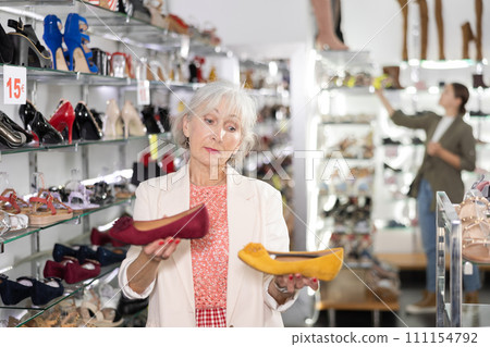 Positive mature customer holds in hands and compares yellow and burgundy ballet flats 111154792
