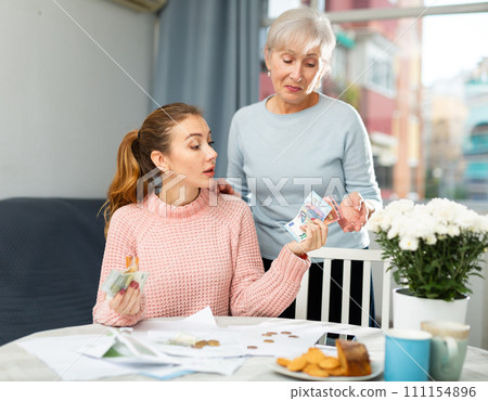 Young woman holding out banknotes to aged mother at home table Young woman holding out banknotes to aged mother at home table 111154896