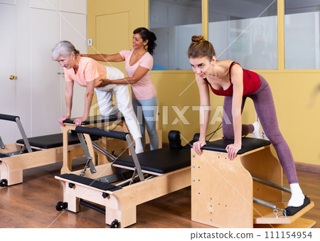 Female instructor helps women perform an exercise on a combined chair 111154954