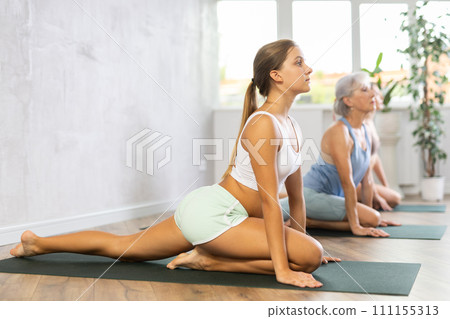 Three generations of women perform sitting pigeon pose Eka Pada Rajakapotasana in gym Three generations of women perform sitting pigeon pose Eka Pada Rajakapotasana in gym 111155313