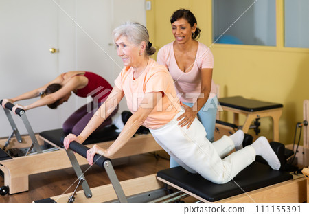 Aged woman doing pilates on reformer with Hispanic female trainer Aged woman doing pilates on reformer with Hispanic female trainer 111155391