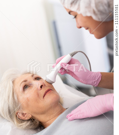 Professional young female cosmetologist doing facial skin ultrasound cavitation for for face lifting of mature woman laying in beauty salon Professional young female cosmetologist doing facial skin ultrasound cavitation for for face lifting of mature woman laying in beauty salon 111155581